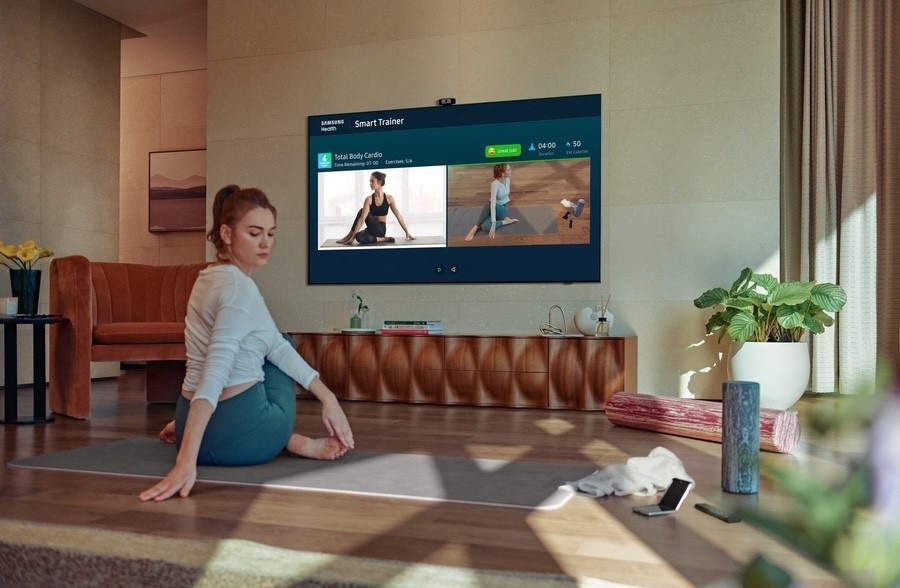 Woman doing yoga at home in front of a Samsung Smart Trainer TV showing a guided total body workout in a stylish living room.