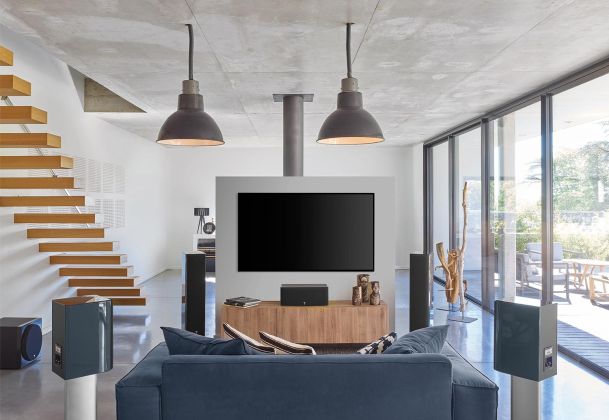 An industrial-style living room with a blue sofa, black speakers, a wall-mounted TV, and large windows overlooking an outdoor deck.