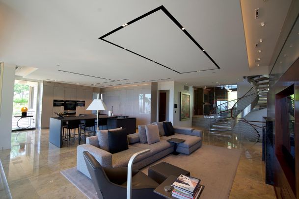 A spacious open-plan living area with a gray sofa, modern kitchen, and a spiral staircase, featuring marble flooring and sleek design elements.